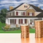 Stacks of coins in front of a large suburban house, symbolizing rising home prices and real estate value.