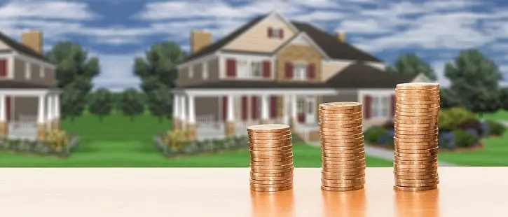 Stacks of coins in front of a large suburban house, symbolizing rising home prices and real estate value.